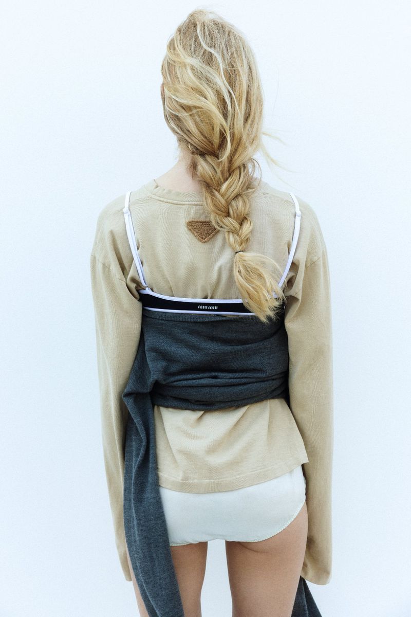 Blonde woman with braided hair faces away from the camera wearing a tan sweater and white bottoms in a minimalistic fashion style.
