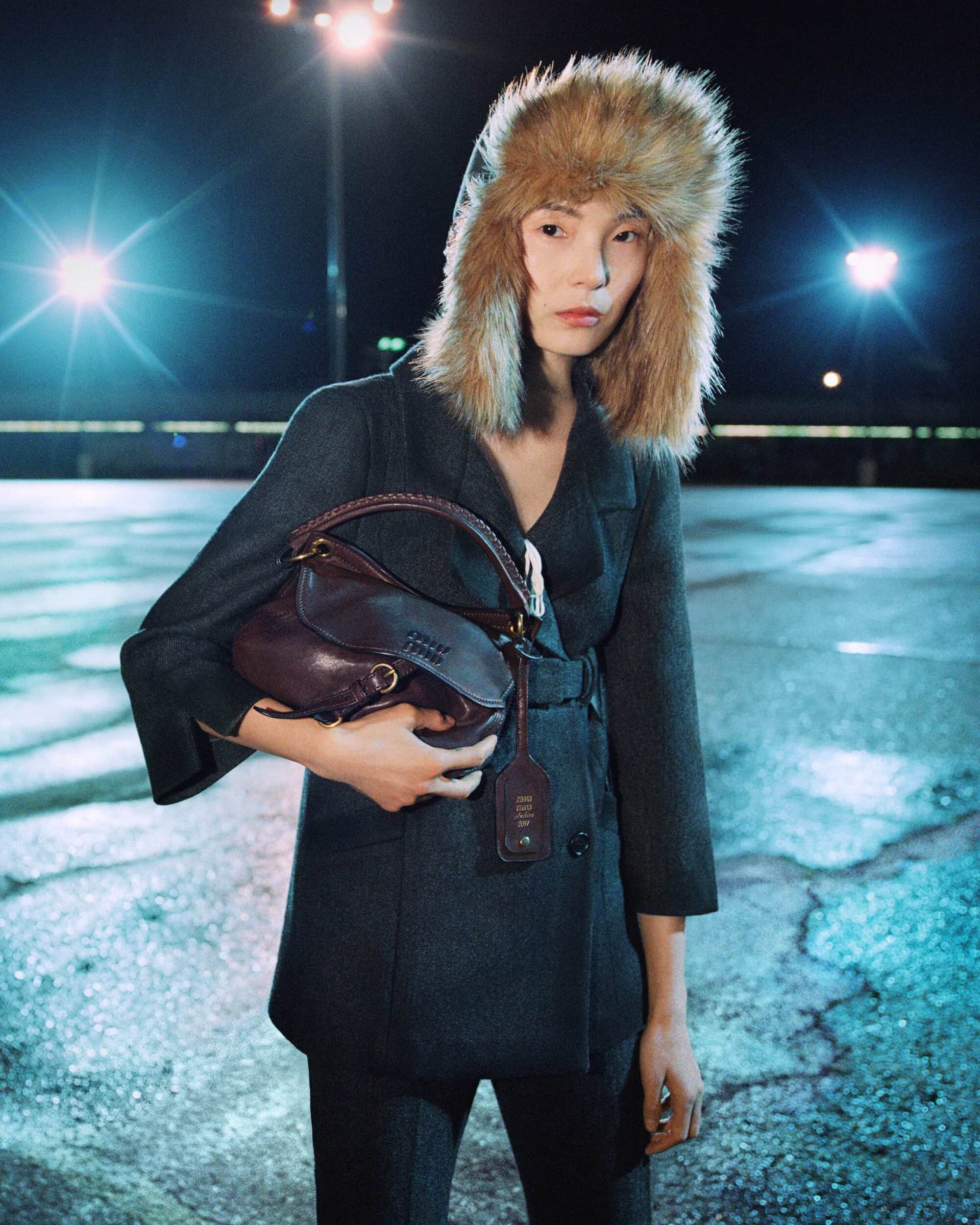 Model Xiao Wen Ju wearing a faux fur trapper hat and charcoal tailored jacket while holding a leather bag in a lit carpark at night.