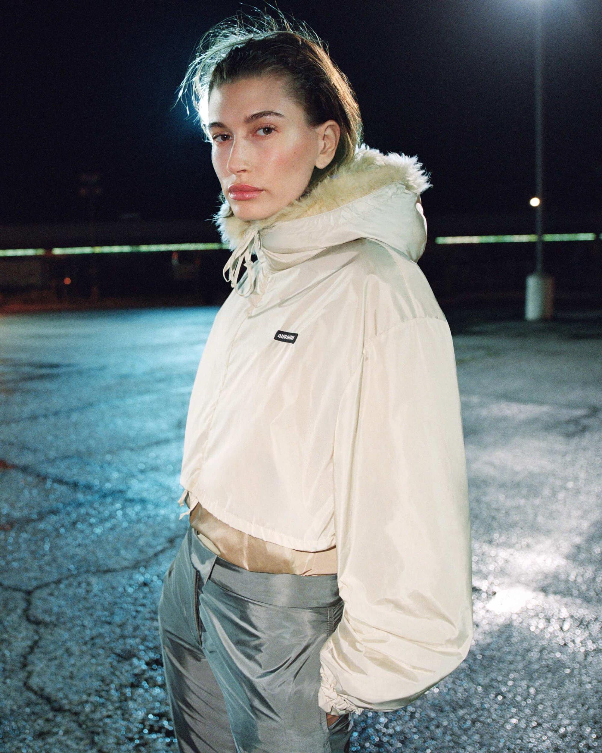 Hailey Bieber wearing a cream hooded jacket and grey trousers outdoors at night under a streetlamp.