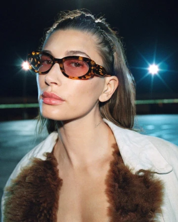 Hailey Bieber wearing tortoiseshell sunglasses with pink tinted lenses and a shearling-trimmed jacket at night.