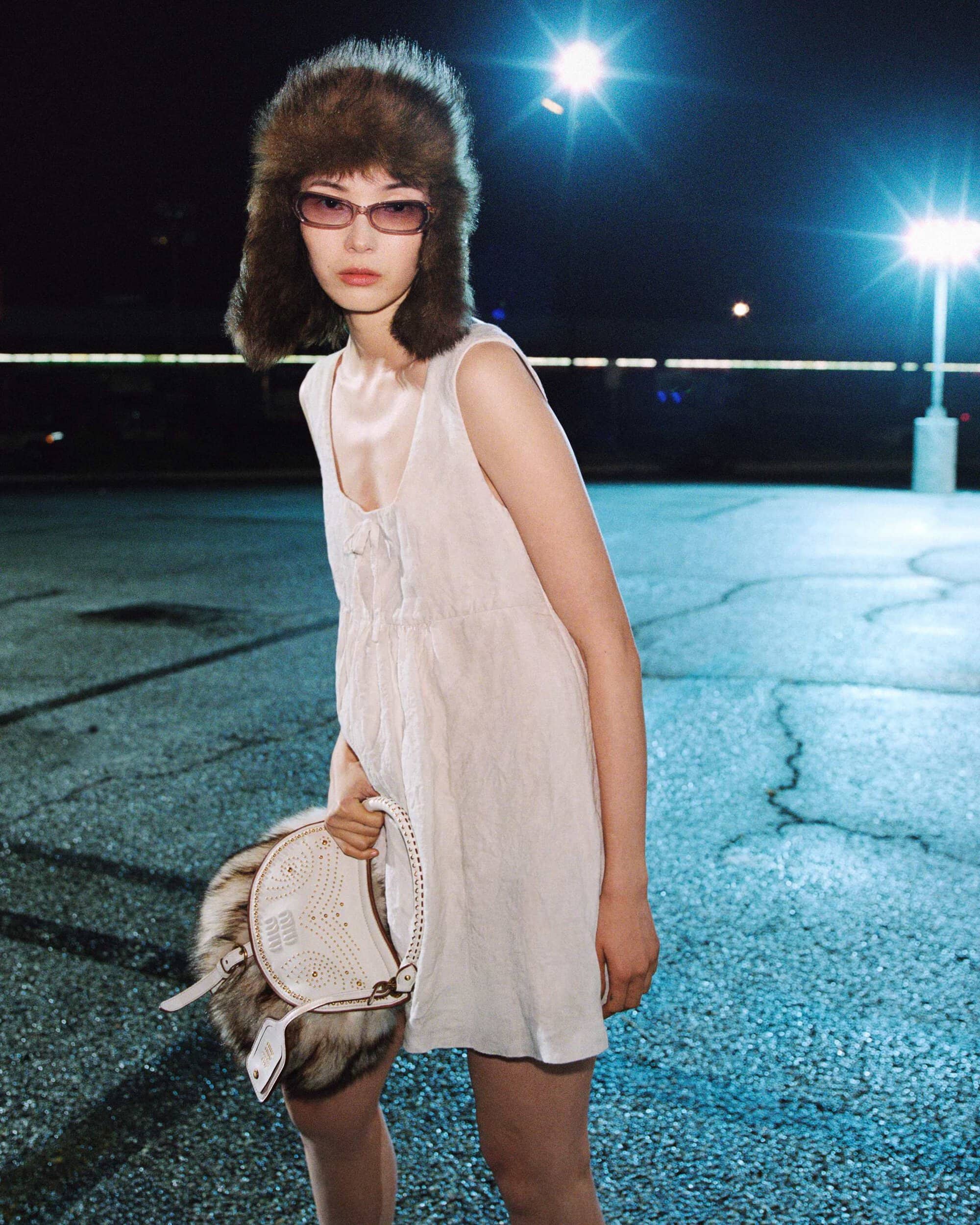 Model Xiao Wen Ju wearing a white slip dress, fur trapper hat, and tinted glasses holding a bag in a parking lot at night.