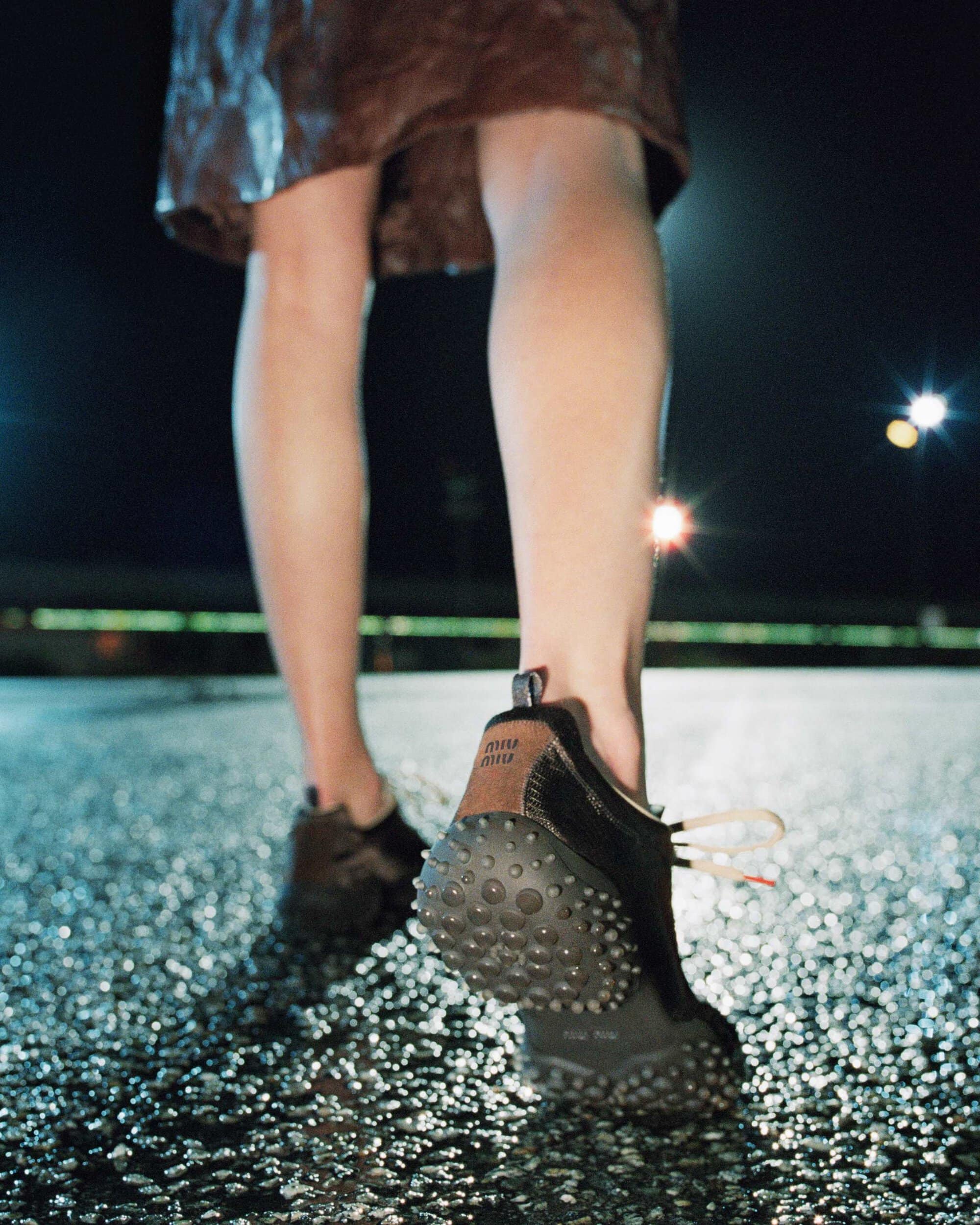 Low-angle rear view of model walking in black Miu Miu sneakers with studded soles on wet pavement at night.