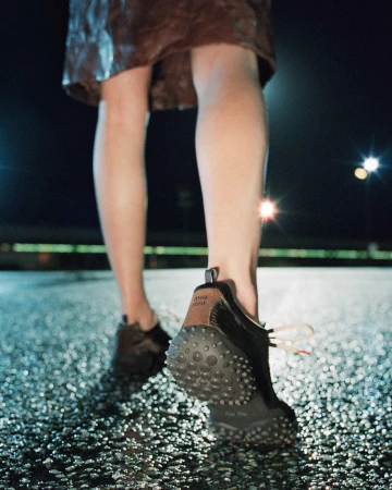 Low-angle rear view of model walking in black Miu Miu sneakers with studded soles on wet pavement at night.