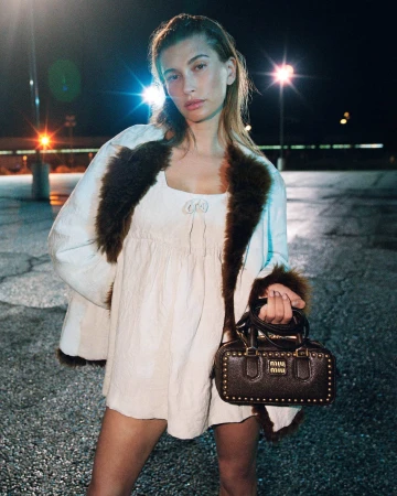 Hailey Bieber wearing a cream mini dress and fur-trimmed coat while holding a studded dark handbag in a parking lot at night.