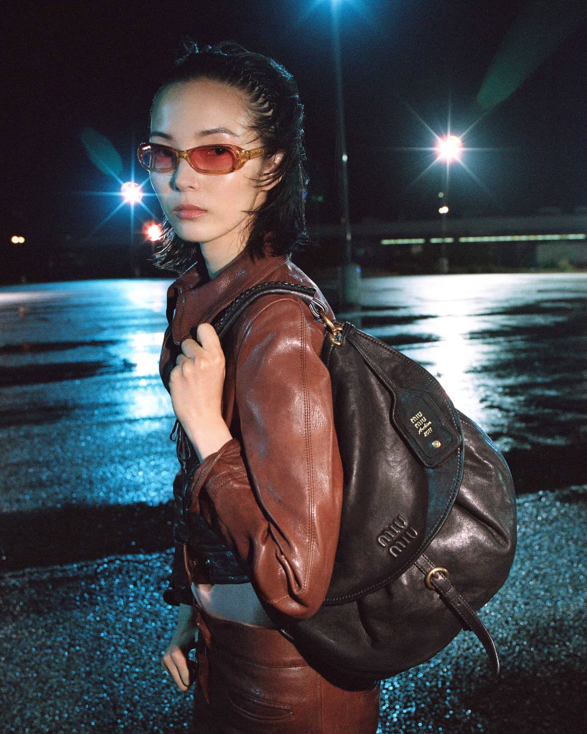 Model Xiao Wen Ju wearing a brown leather jacket and tinted sunglasses with a black Miu Miu shoulder bag at night.