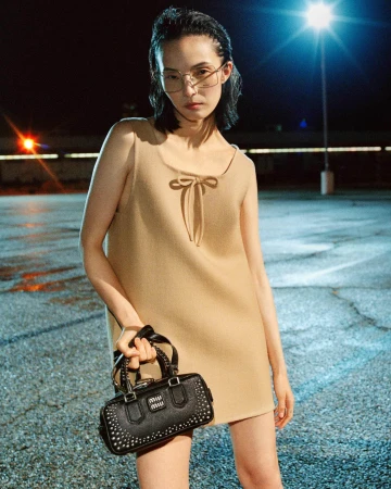 Model Xiao Wen Ju wearing a beige mini dress holding a black studded handbag outdoors at night under a streetlamp for Miu Miu.
