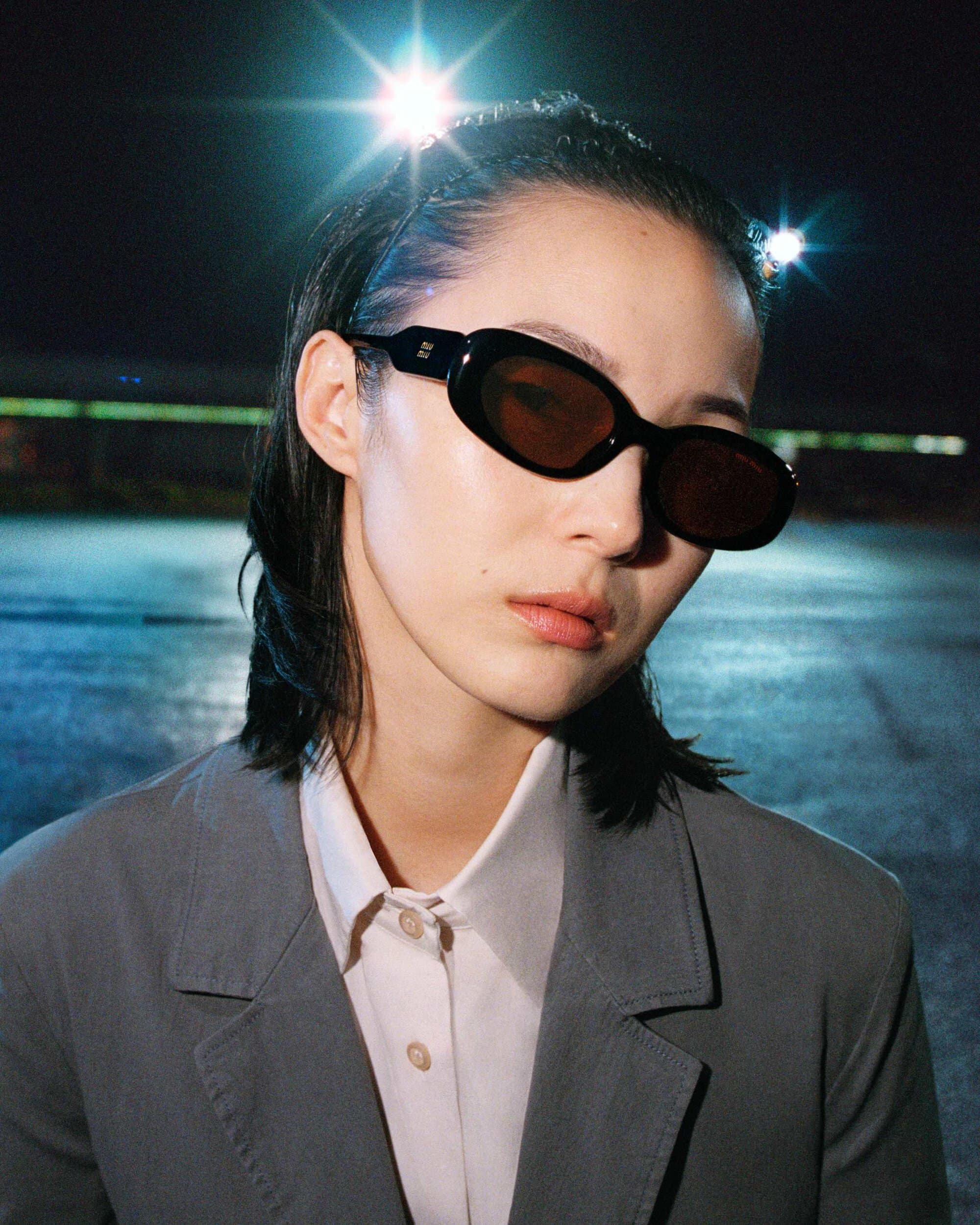 Xiao Wen Ju wearing black oval sunglasses, a white collared shirt, and a grey blazer under night floodlights.