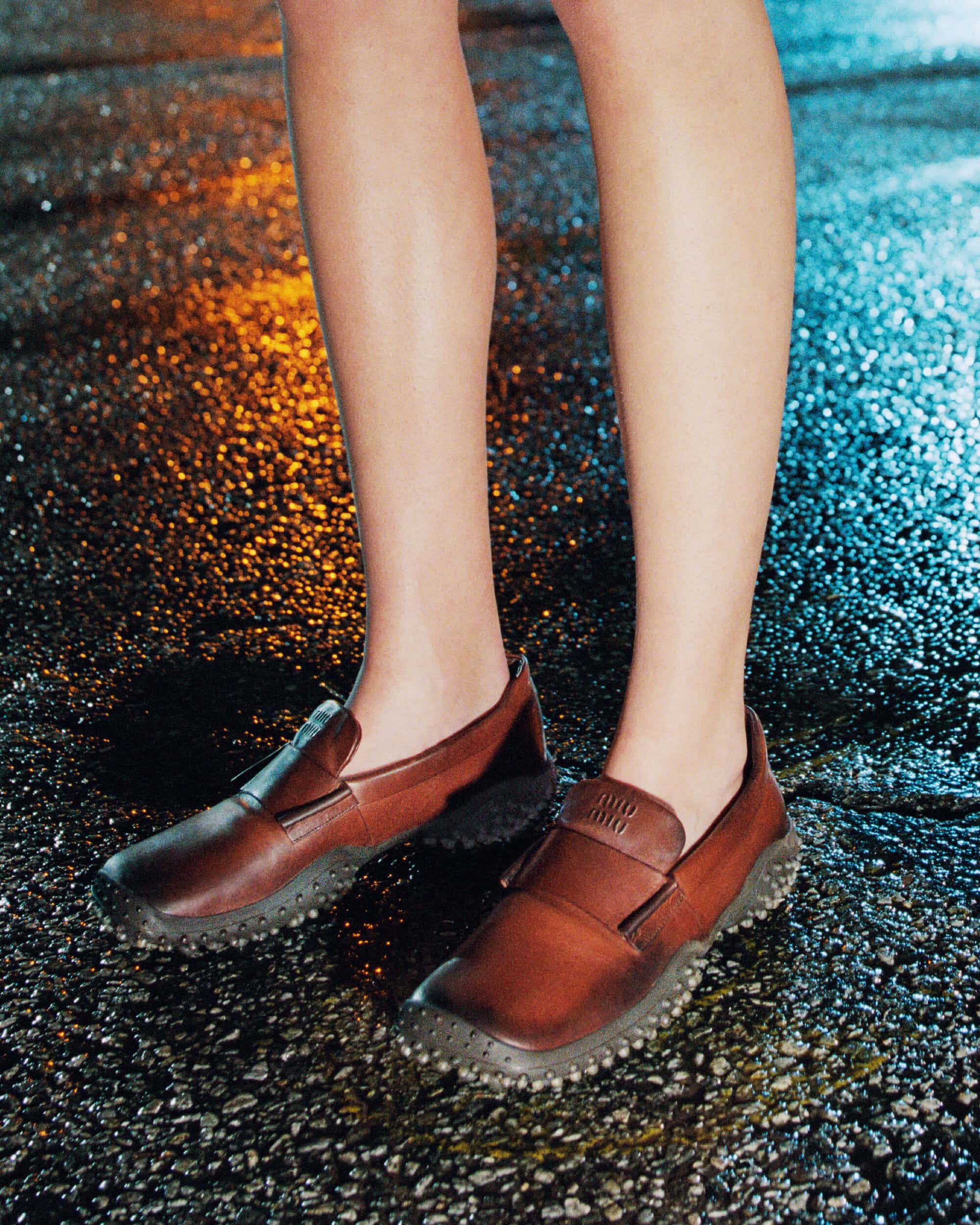 Close-up of bare legs wearing brown leather slip-on loafers standing on wet asphalt with colored light reflections.