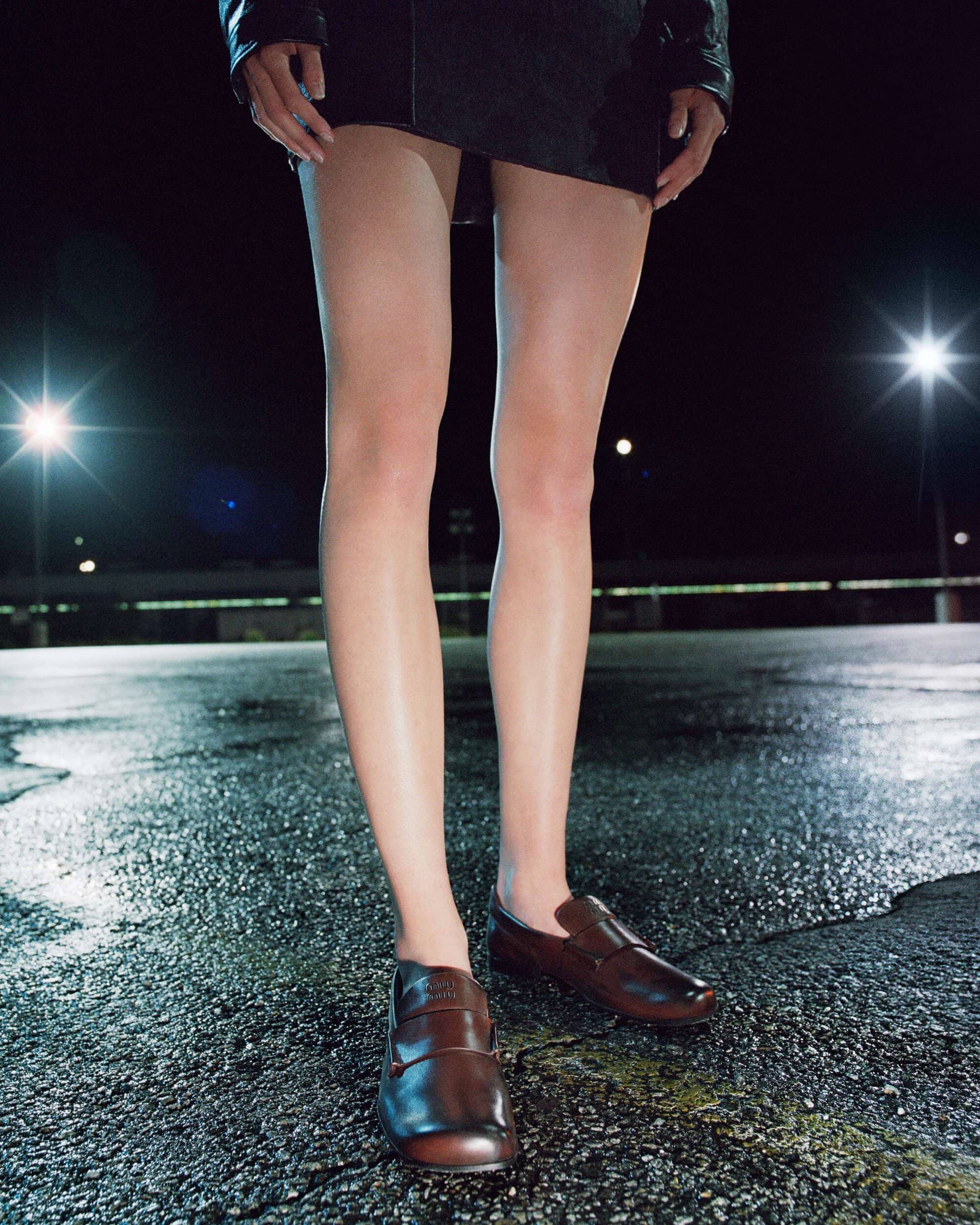 Close-up of model's legs wearing brown leather loafers and a black mini skirt on wet asphalt at night under bright lights.