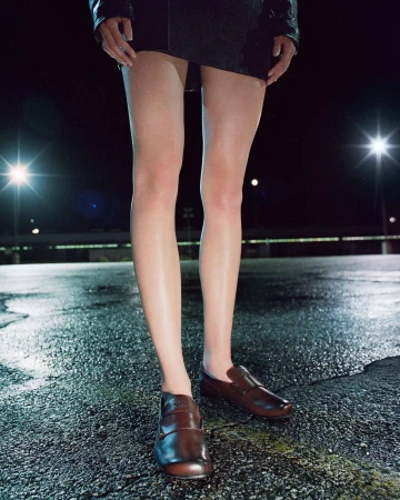 Close-up of model's legs wearing brown leather loafers and a black mini skirt on wet asphalt at night under bright lights.