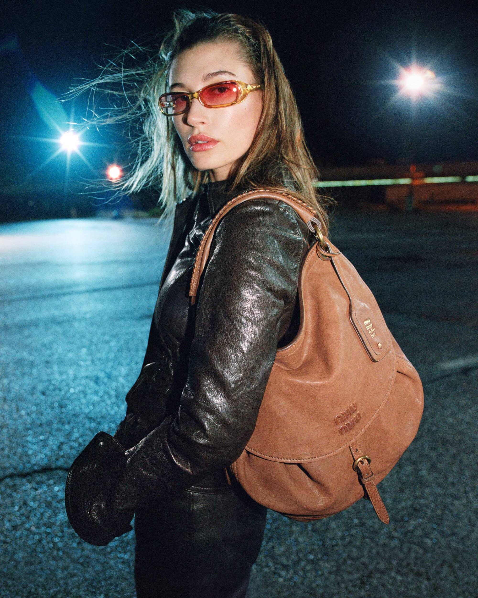Hailey Bieber wearing tinted sunglasses, a black leather jacket, and carrying a tan leather shoulder bag outdoors at night.