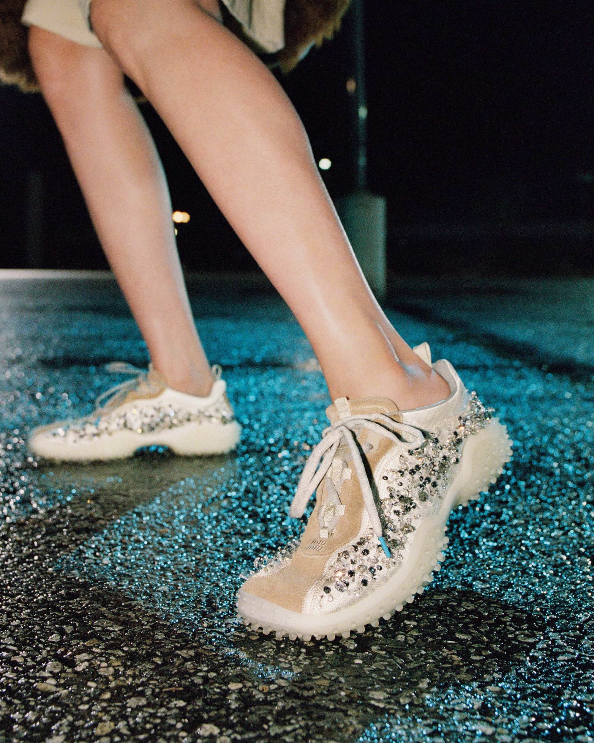 Close-up of bare legs wearing crystal-embellished cream sneakers with textured rubber soles standing on wet asphalt at night.
