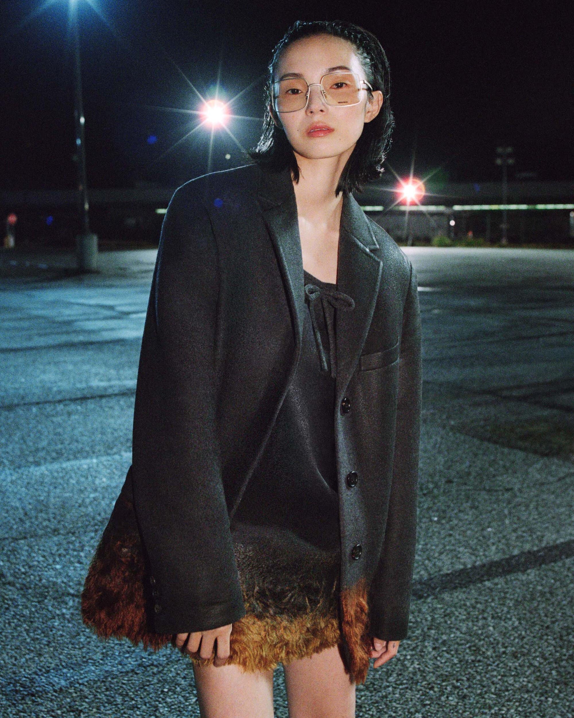 Model Xiao Wen Ju wearing a dark tailored blazer and tinted eyewear in an open asphalt parking lot at night under floodlights.