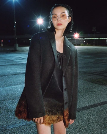 Model Xiao Wen Ju wearing a dark tailored blazer and tinted eyewear in an open asphalt parking lot at night under floodlights.