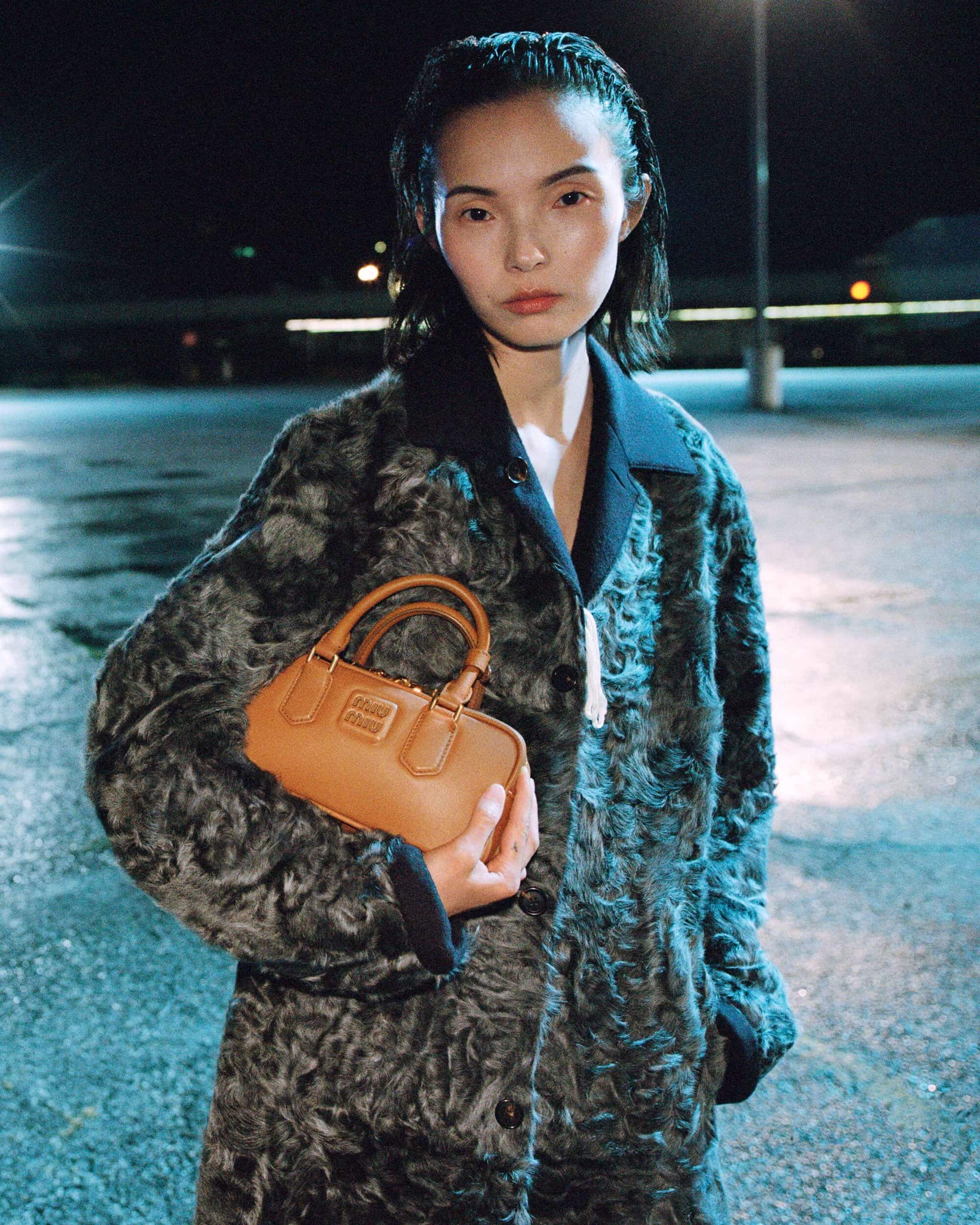 Model Xiao Wen Ju wearing a textured grey fur coat and holding a tan leather mini handbag in a parking lot at night.
