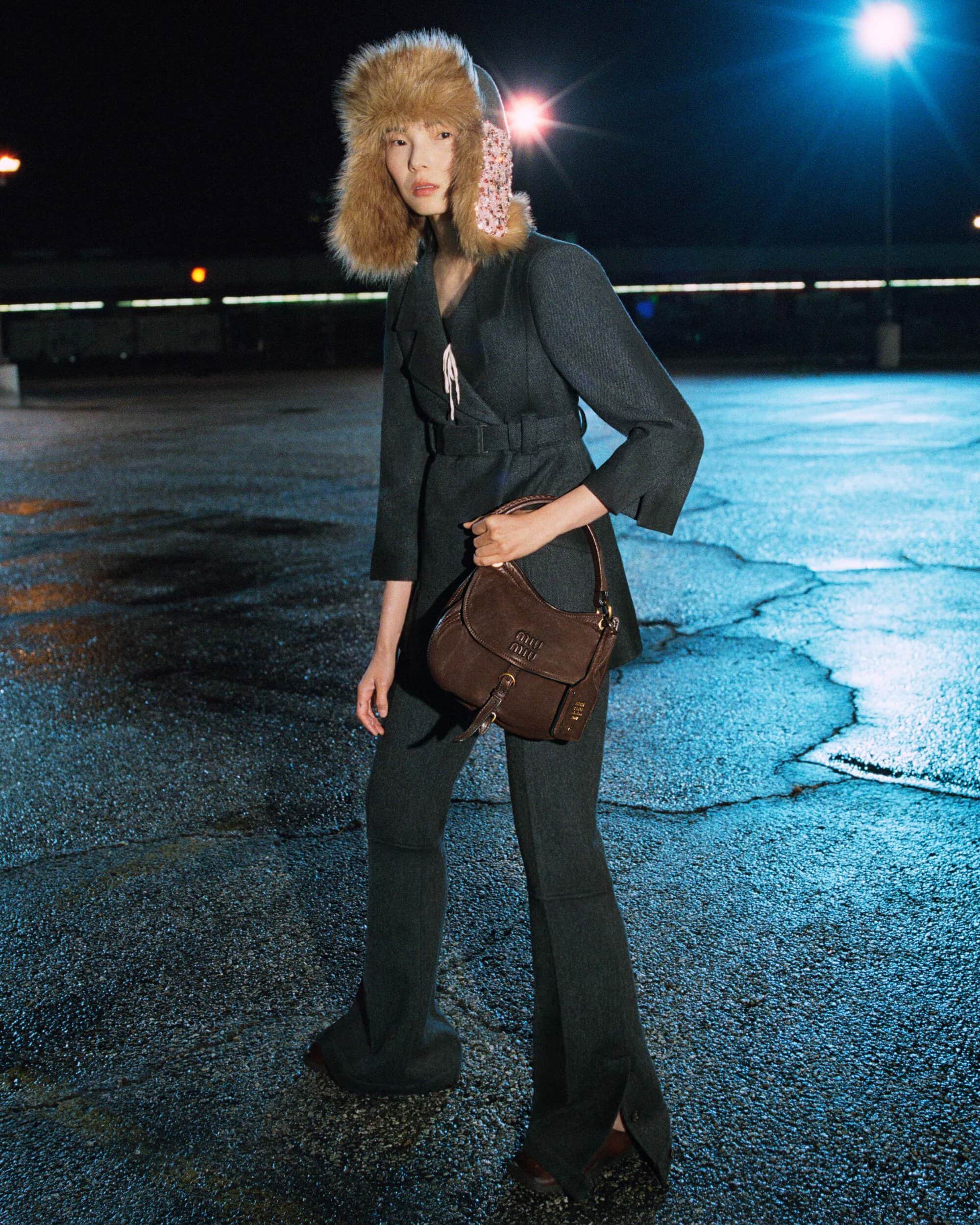 Xiao Wen Ju wearing a charcoal belted suit, a fur trapper hat, and holding a brown leather bag on wet asphalt at night.