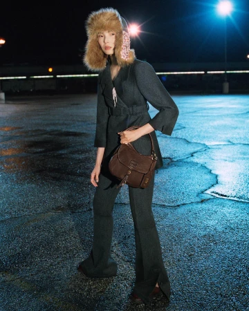 Xiao Wen Ju wearing a charcoal belted suit, a fur trapper hat, and holding a brown leather bag on wet asphalt at night.