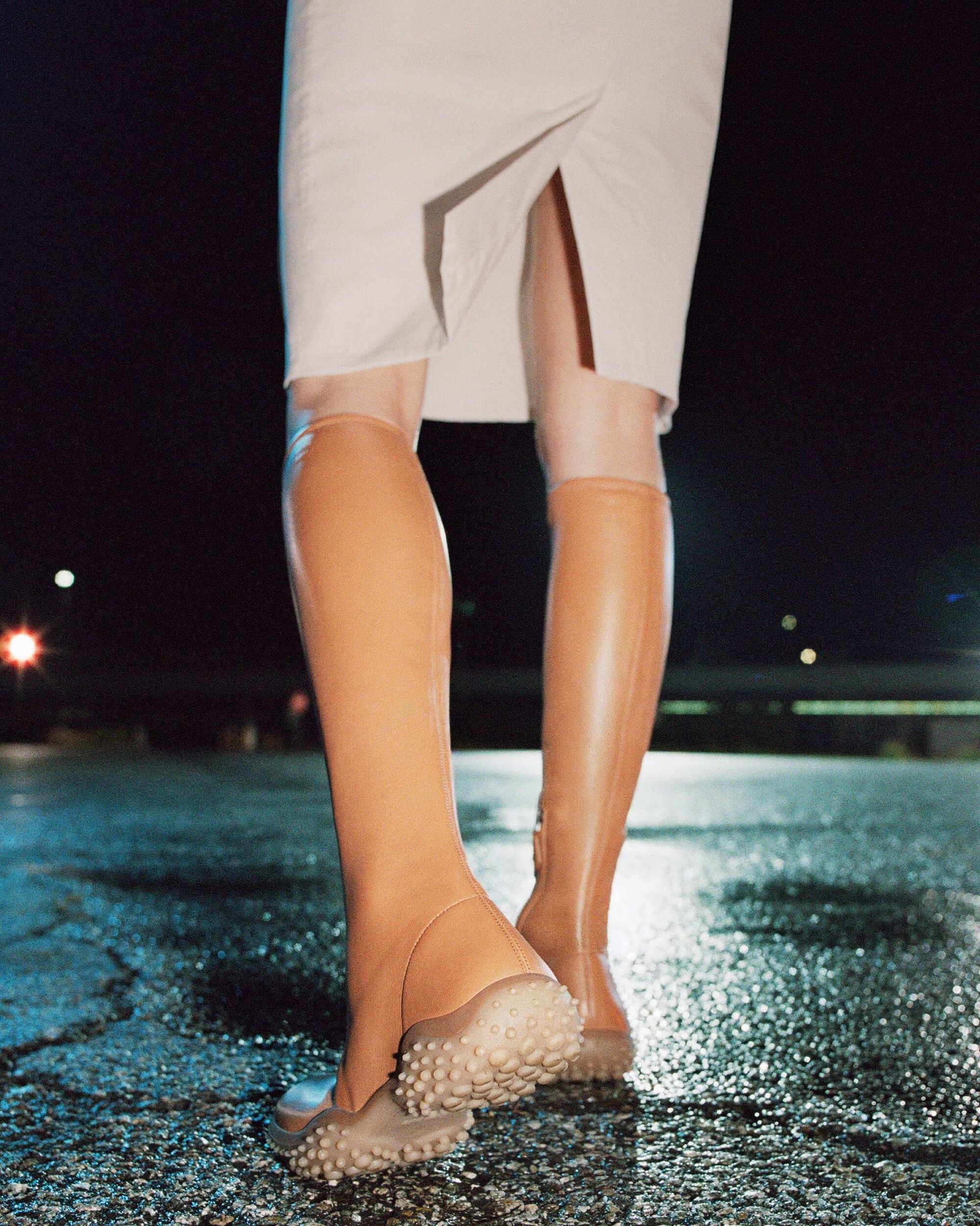 Close-up rear view of model wearing camel knee-high boots with pebbled soles and a white slit skirt on wet asphalt at night.