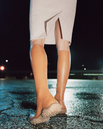 Close-up rear view of model wearing camel knee-high boots with pebbled soles and a white slit skirt on wet asphalt at night.