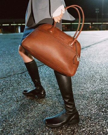 Cropped view of a model holding a large brown leather Miu Miu handbag while wearing black knee-high leather boots on asphalt.