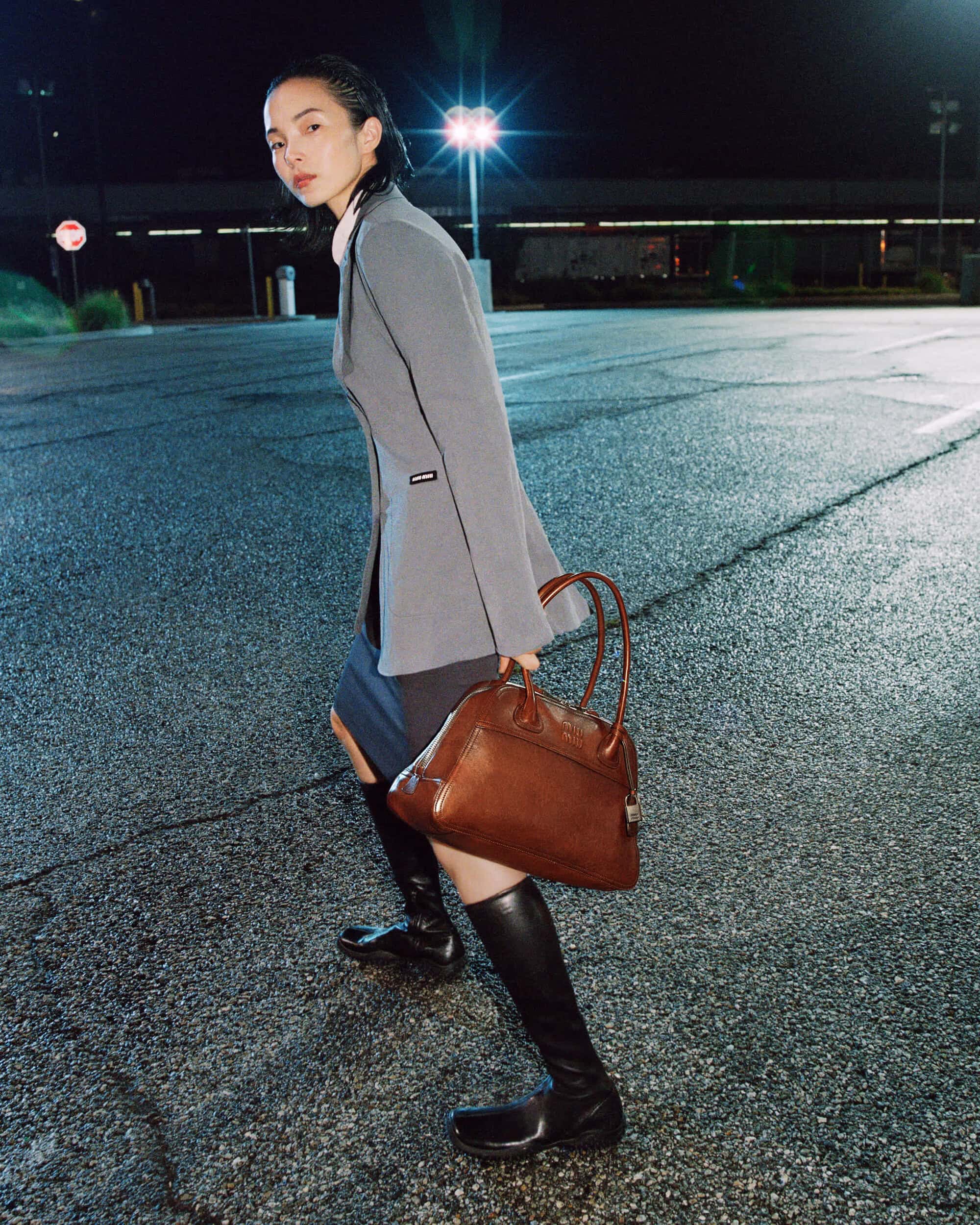 Xiao Wen Ju carries a brown leather handbag while standing on an asphalt parking lot at night in a grey jacket and knee-high boots.