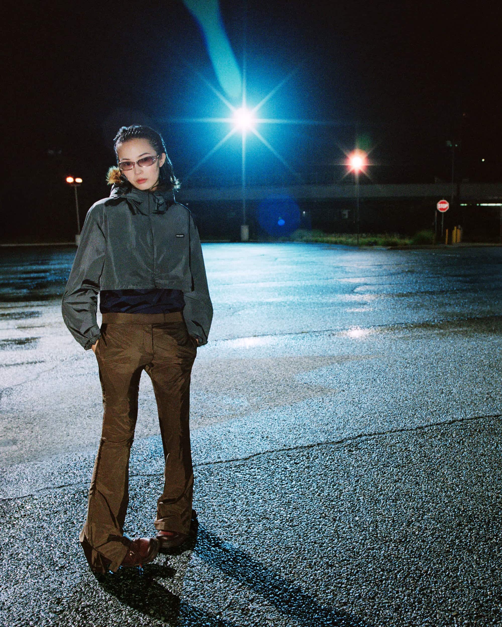 Model Xiao Wen Ju wearing a grey hooded windbreaker and brown flared pants in a dark parking lot for Miu Miu.