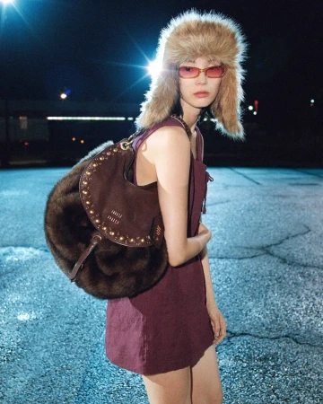Model Xiao Wen Ju wearing a furry ushanka hat, tinted sunglasses, and a plum mini dress while carrying a brown fur shoulder bag.