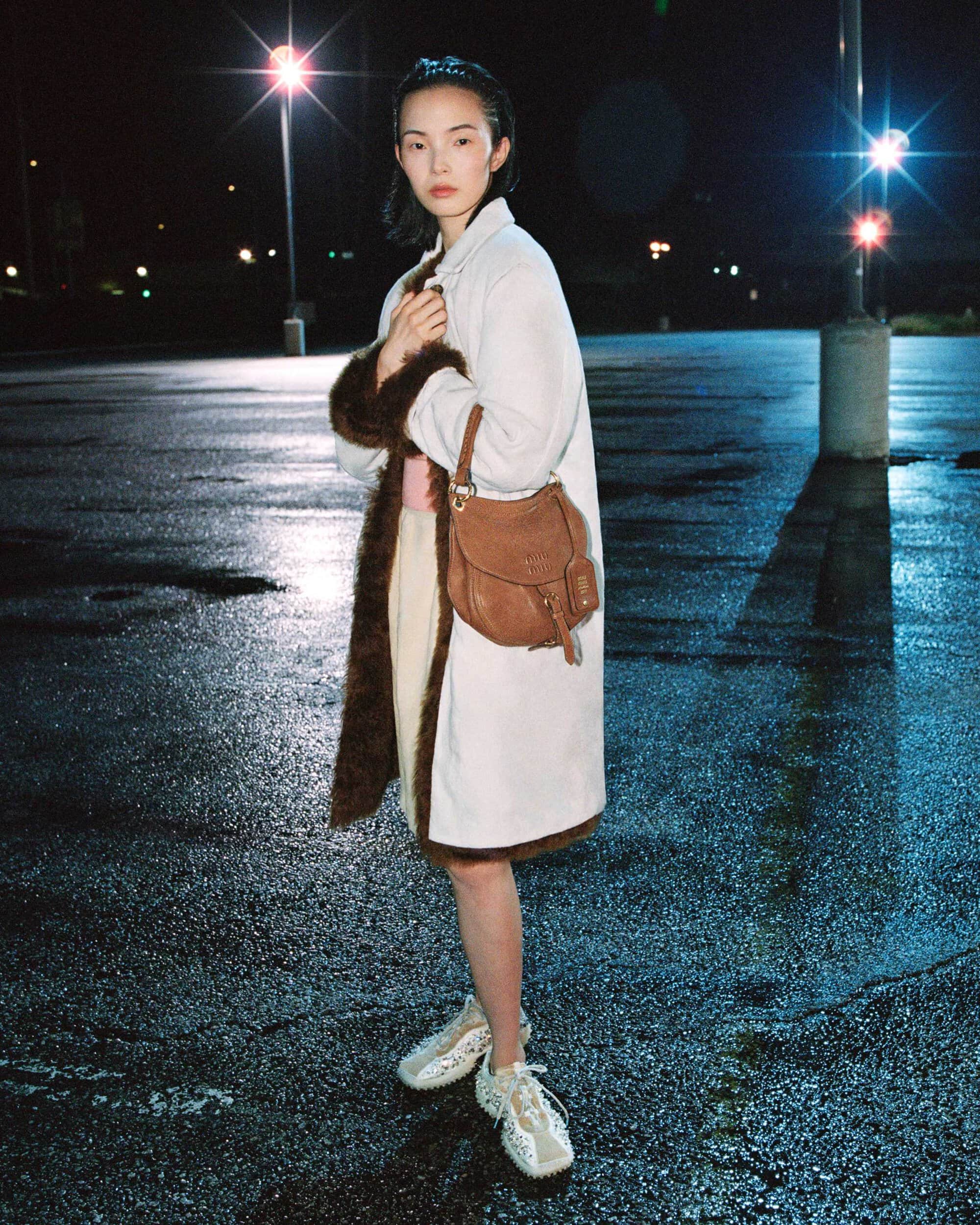 Model Xiao Wen Ju wearing a white fur-trimmed coat and brown leather shoulder bag in a wet parking lot at night.