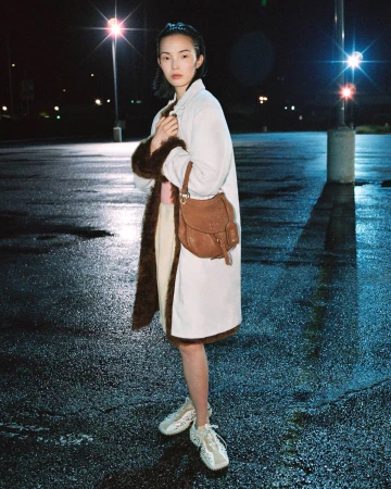 Model Xiao Wen Ju wearing a white fur-trimmed coat and brown leather shoulder bag in a wet parking lot at night.