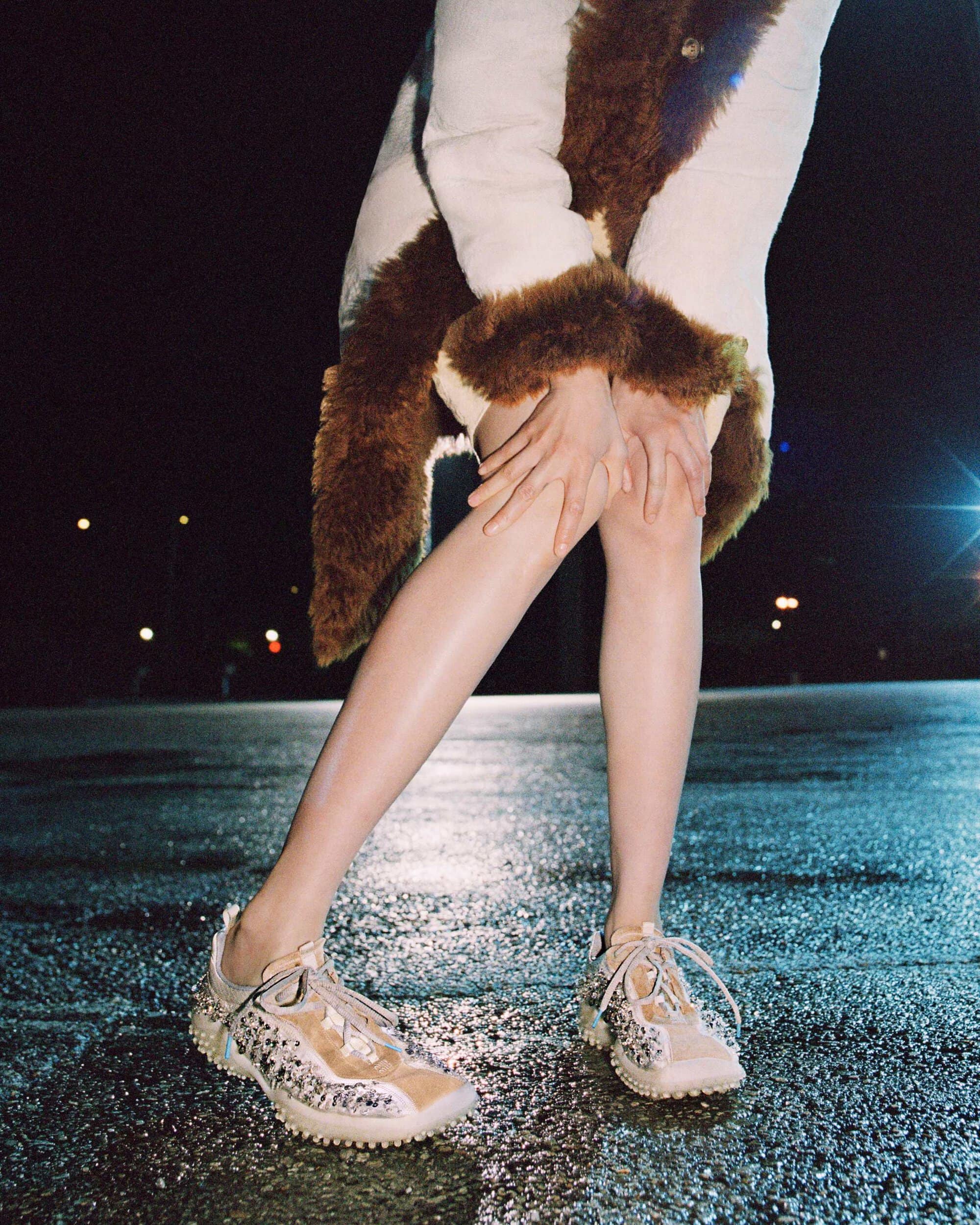 Cropped view of a model leaning forward in a brown fur-trimmed coat and low-top sneakers on wet asphalt at night.