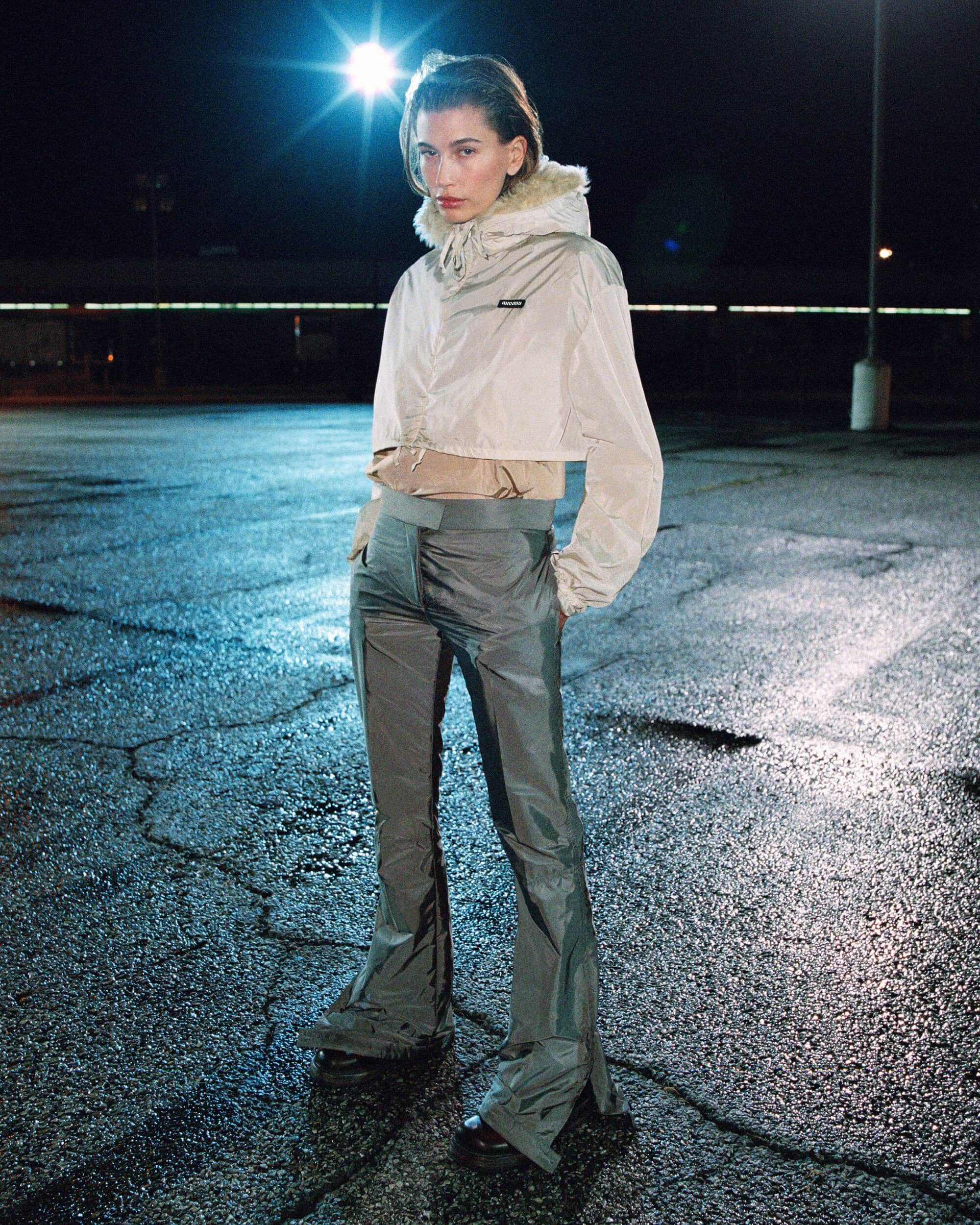 Hailey Bieber stands outdoors at night on asphalt wearing a cropped hooded beige jacket and grey flared trousers.