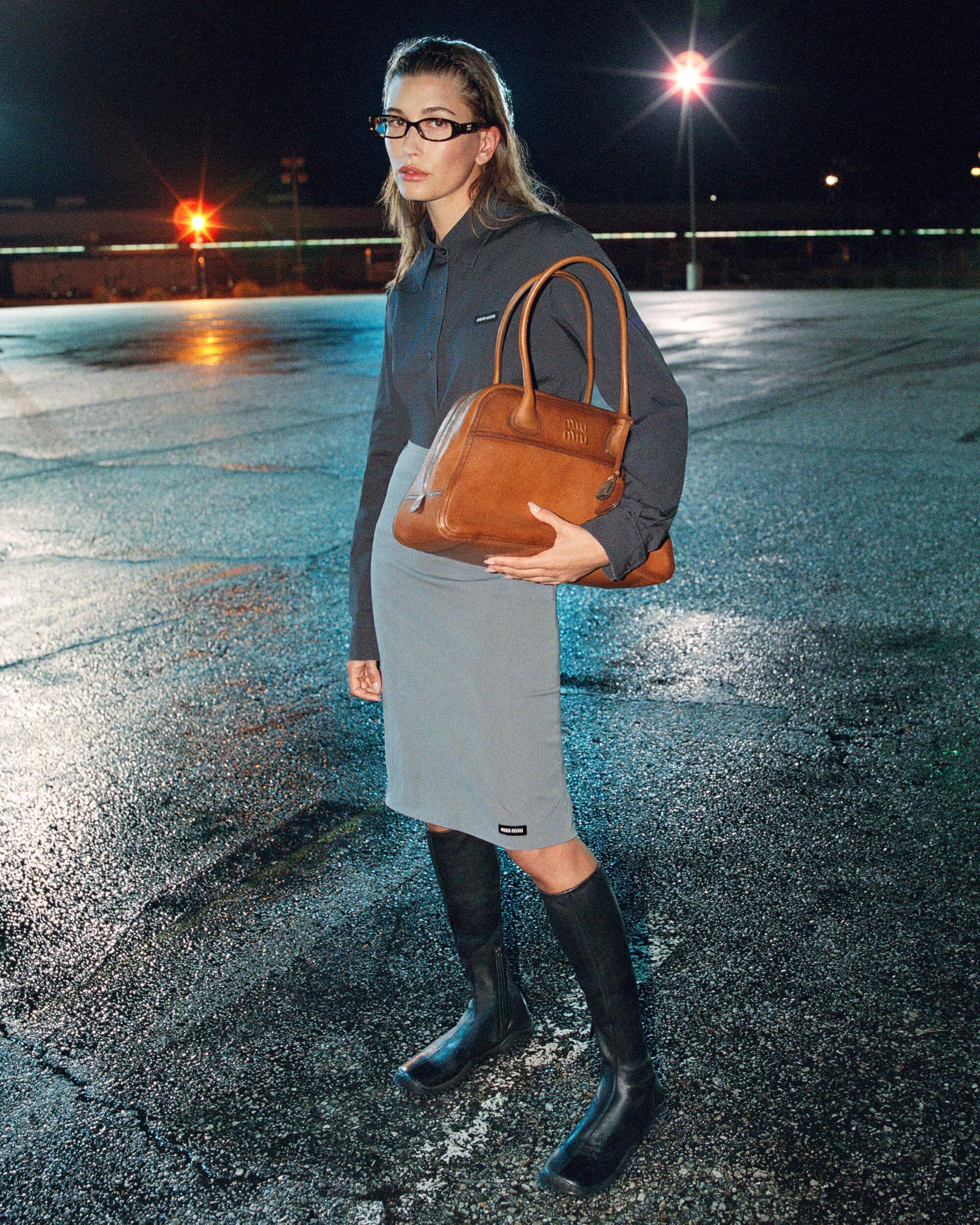 Hailey Bieber wearing glasses and holding a tan leather bag outdoors at night on a wet asphalt surface for Miu Miu