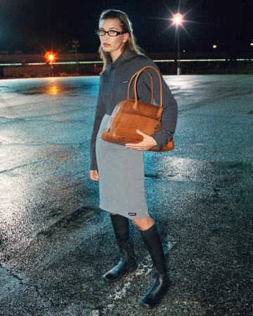 Hailey Bieber wearing glasses and holding a tan leather bag outdoors at night on a wet asphalt surface for Miu Miu