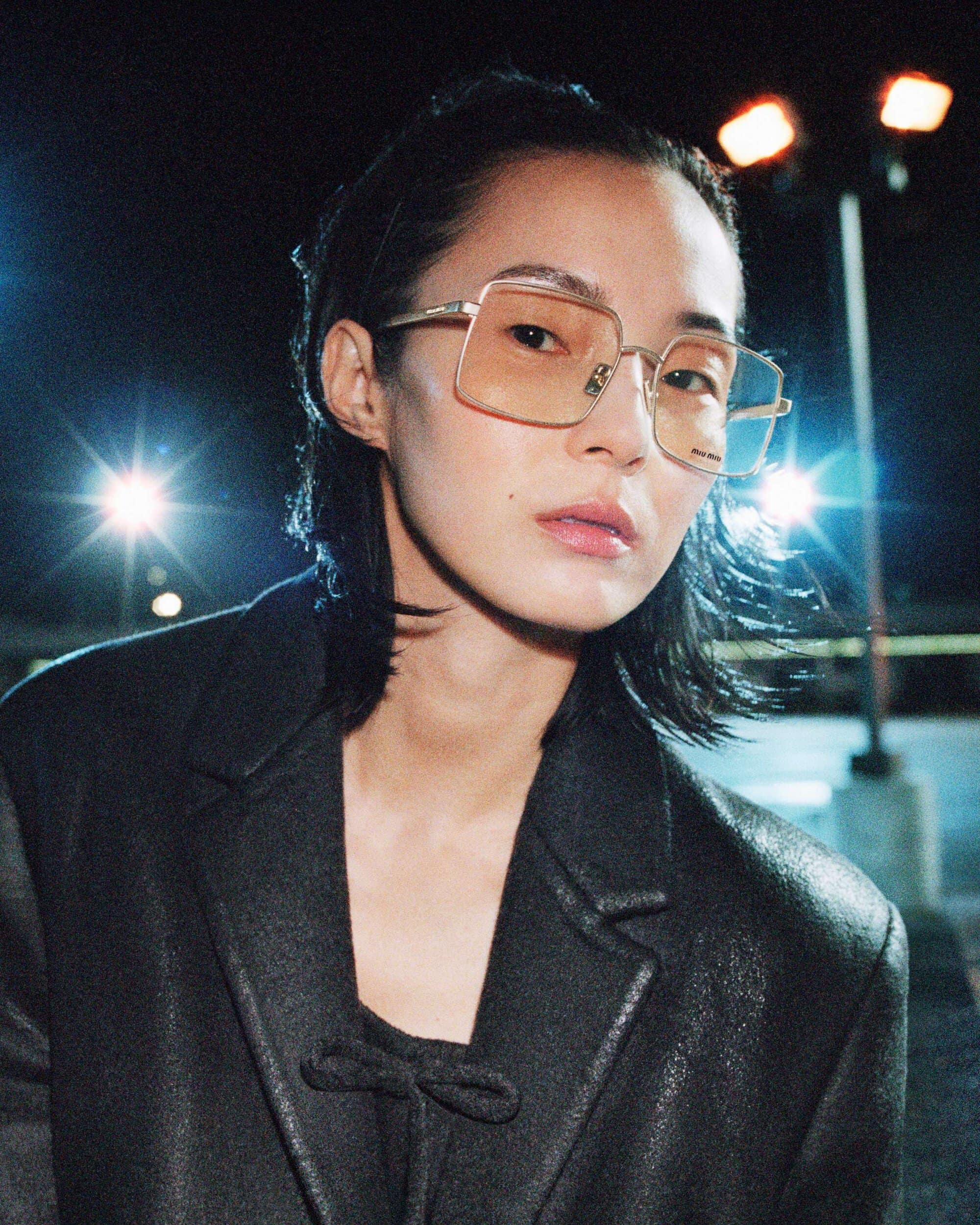 Xiao Wen Ju wearing oversized square wireframe tinted glasses and a black textured coat against nighttime lights.