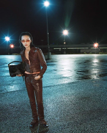 Model Xiao Wen Ju posing in a brown leather jacket, matching pants, and tinted sunglasses while holding a black handbag in a parking lot.
