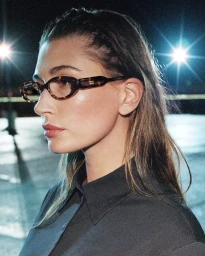 Hailey Bieber wearing tortoiseshell oval optical glasses and a dark grey collared shirt in profile against bright night lights.
