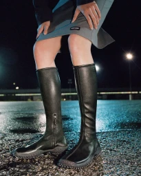 Close-up of a model wearing black knee-high leather boots with rubber soles and a grey skirt on wet asphalt at night.