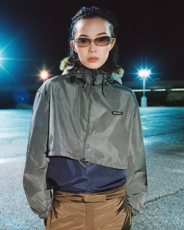 Model wearing tinted sunglasses, a cropped grey hooded windbreaker, and brown trousers in an asphalt lot at night.