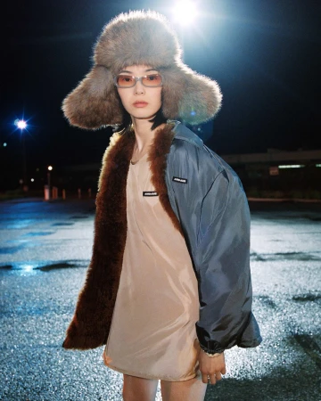 Xiao Wen Ju wearing an oversized brown fur ushanka hat, tinted glasses, and a fur-trimmed jacket in a wet carpark at night.