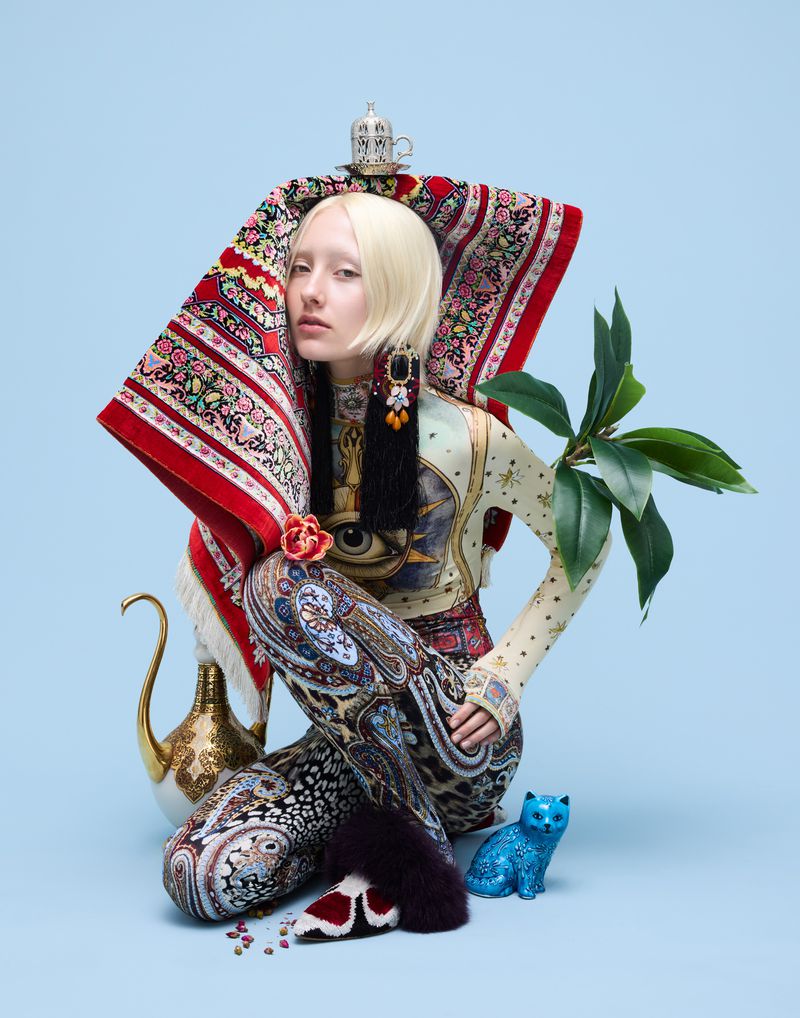 Woman with pale skin and platinum blonde hair poses with decorative rug, tea pot, and blue cat figure on a light blue background.
