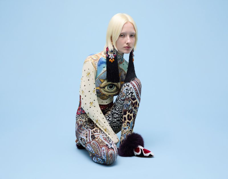 Woman in patterned bodysuit and tasseled earrings squats in front of a light blue backdrop in a fashion editorial studio shot.
