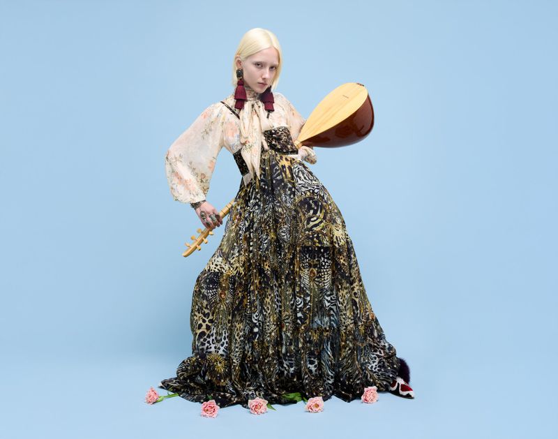 Woman posing with stringed instruments against a light blue backdrop, wearing a long patterned dress, earrings, and floral accents.