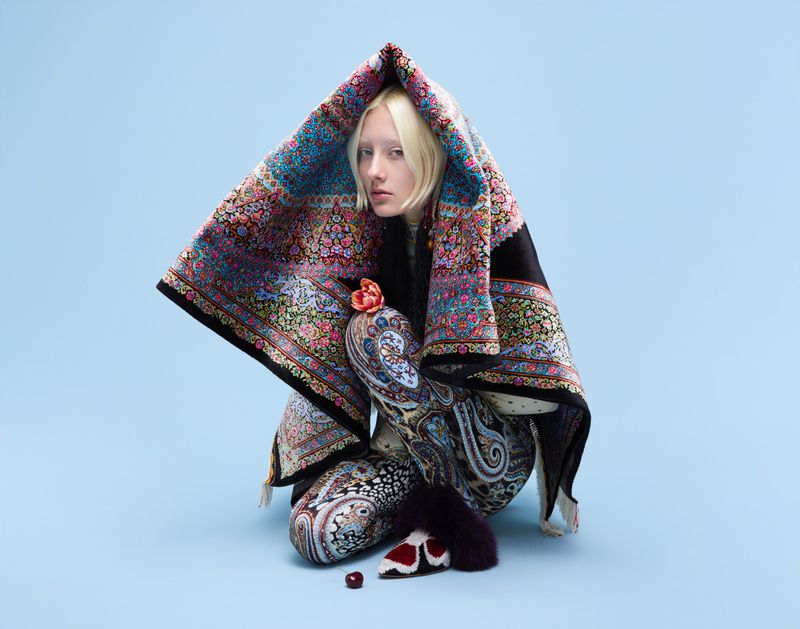 Young woman with fair skin and blond hair wearing patterned hooded cloak and leggings against a light blue backdrop in a studio shot.