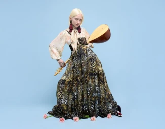 Blonde model in a patterned Camilla maxi dress holding a wooden lute against a light blue studio background with pink roses at her feet.