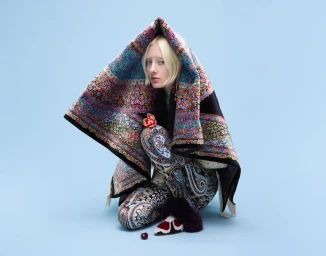 Model with blonde hair crouching under a patterned blanket against a light blue background holding a flower