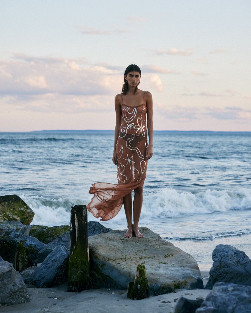 Model Rossana Latallada standing barefoot on rocks by the ocean in a sheer brown patterned slip dress for Alix of Bohemia.