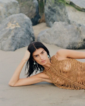 Model Rossana Latallada lying on a sandy beach wearing an earthy brown beaded halter top against coastal rocks.