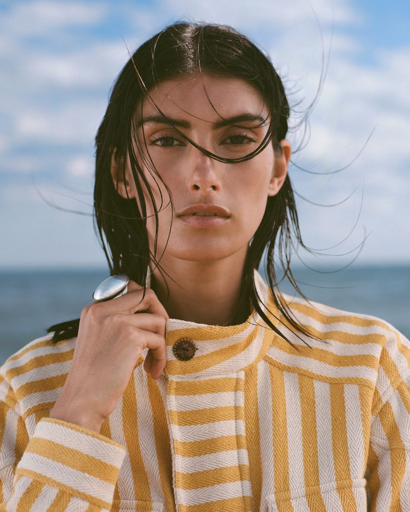Model Rossana Latallada wearing a yellow and white striped jacket and a silver ring outdoors against the sea.