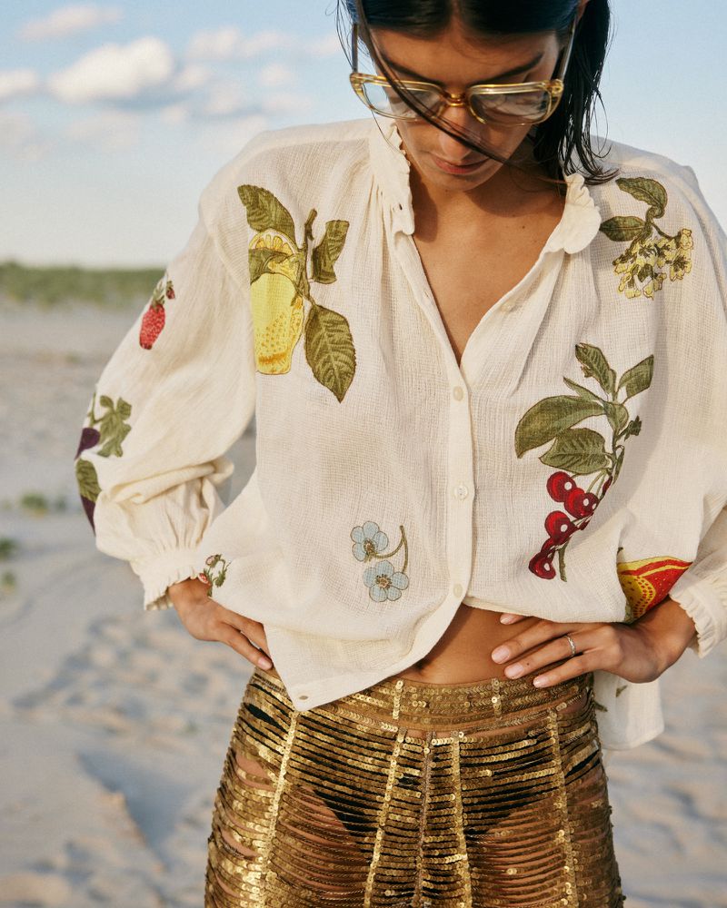Model Rossana Latallada wearing an embroidered fruit-motif white blouse and gold sequin pants on a sandy beach.