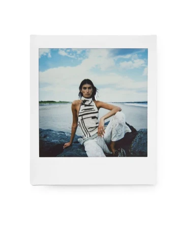 Model wearing patterned halter top and white trousers seated on coastal rocks against a sandy beach and ocean backdrop.